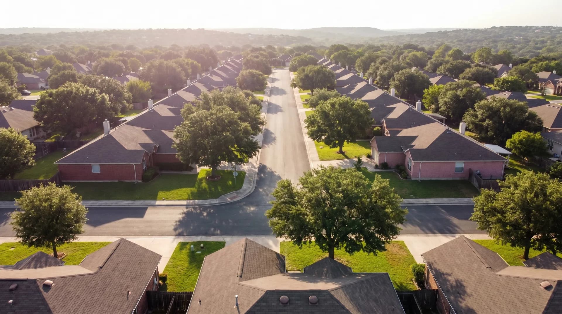 Aerial view of a San Antonio suburban subdivision at golden hour: rows of single-story brick ranch homes with composition-shingle roofs line a straight residential street, interspersed with mature live oak trees and green lawns, with Hill Country tree cover on the horizon.